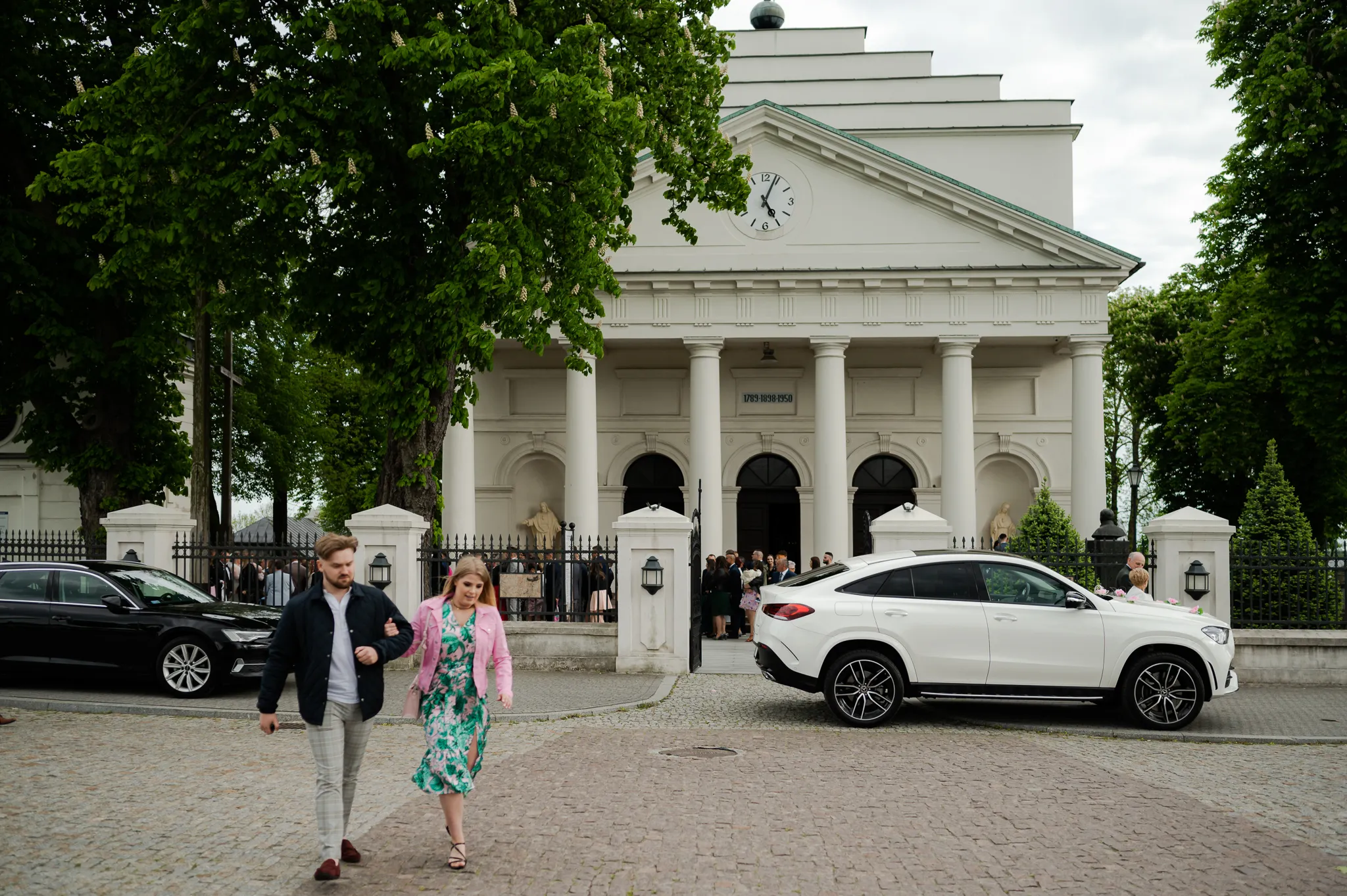 Widok zewnętrzny kościoła w dniu ślubu z klasyczną architekturą i kolumnami, goście weselni w eleganckich strojach, samochody zaparkowane przed świątynią