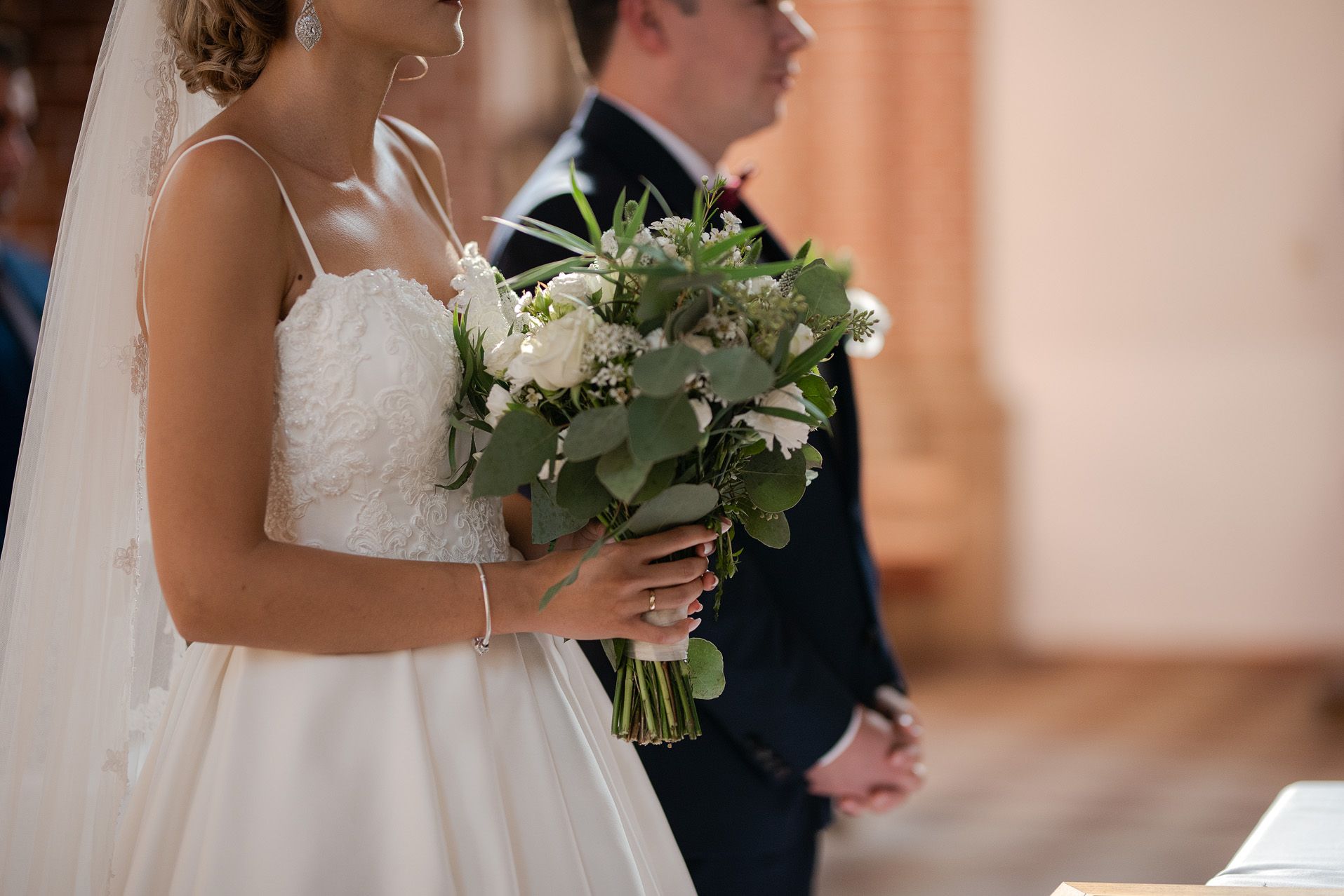 Zbliżenie na bukiet ślubny z białych róż i zieleni trzymany przez pannę młodą w koronkowej sukni podczas ceremonii kościelnej