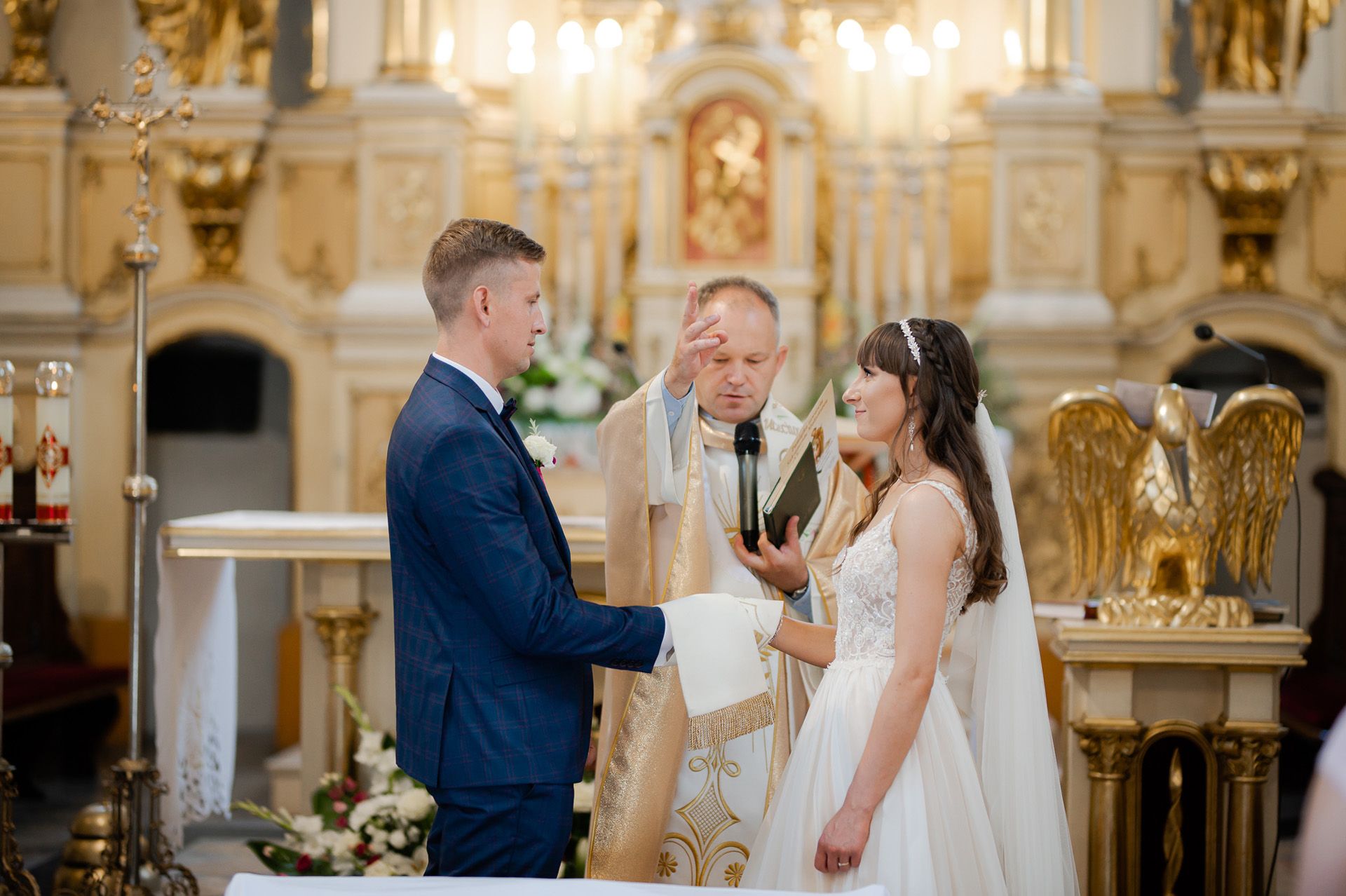Moment błogosławieństwa podczas ceremonii ślubnej przy bogato zdobionym złotym ołtarzu - ksiądz udziela sakramentu, para młoda w białej sukni i granatowym garniturze