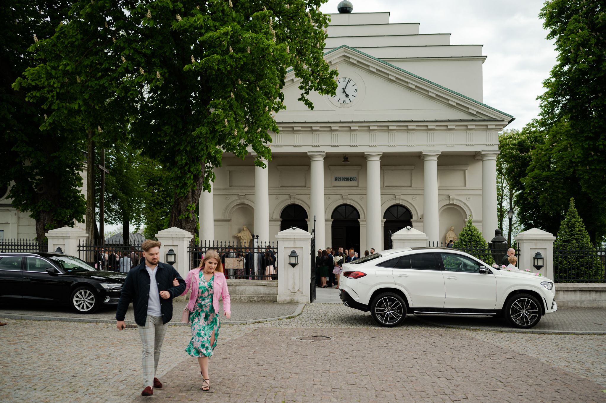 Widok zewnętrzny kościoła w dniu ślubu z klasyczną architekturą i kolumnami, goście weselni w eleganckich strojach, samochody zaparkowane przed świątynią