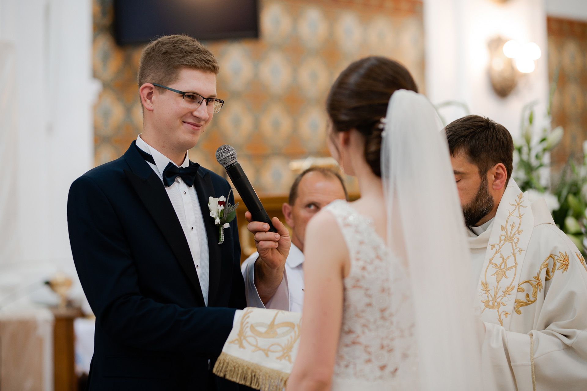 Pan młody w okularach i czarnym garniturze z muszką wypowiada przysięgę ślubną przez mikrofon podczas ceremonii kościelnej