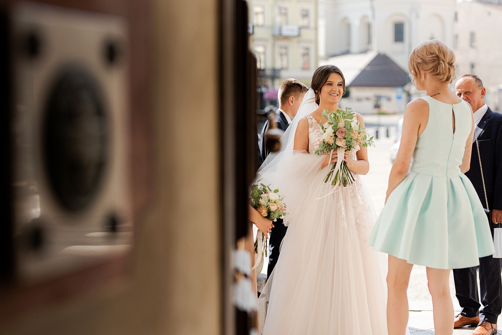 Panna młoda w tiulowej sukni ślubnej z bukietem przybywająca do kościoła, goście w tle, moment przed ceremonią ślubną