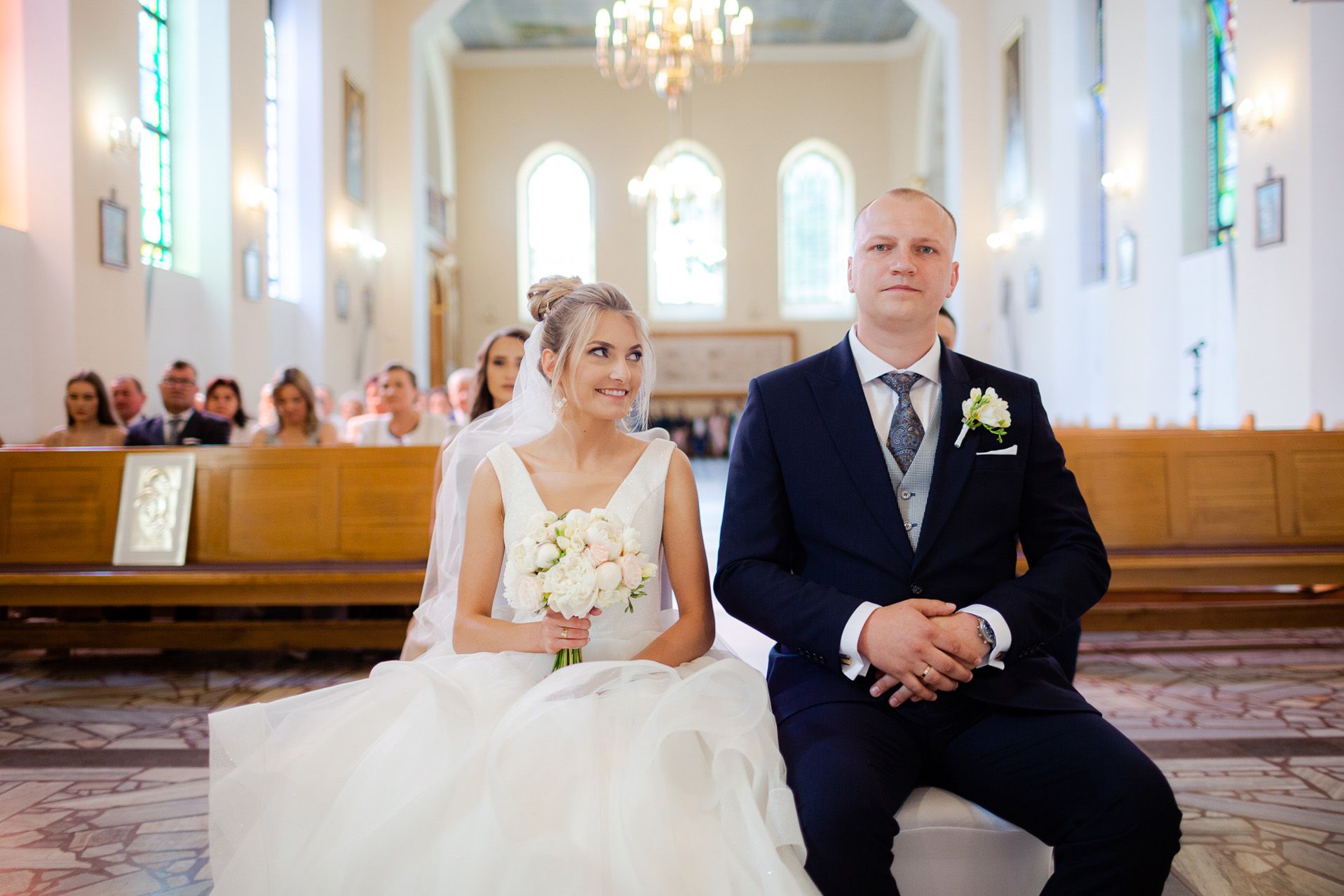 Para młoda siedzi w ławce kościelnej podczas ceremonii ślubnej - panna młoda w białej sukni z bukietem białych kwiatów, pan młody w granatowym garniturze