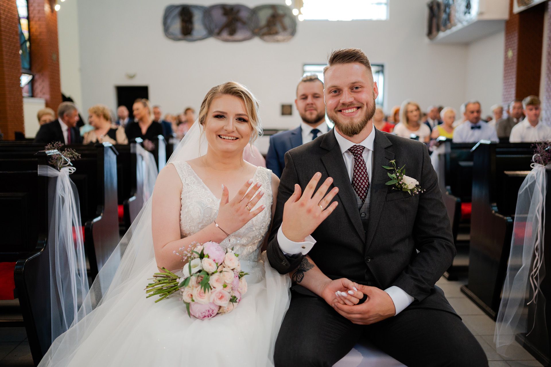 Para młoda pokazuje obrączki ślubne z uśmiechem siedząc w ławce kościelnej, panna młoda w białej sukni z bukietem, pan młody w szarym garniturze