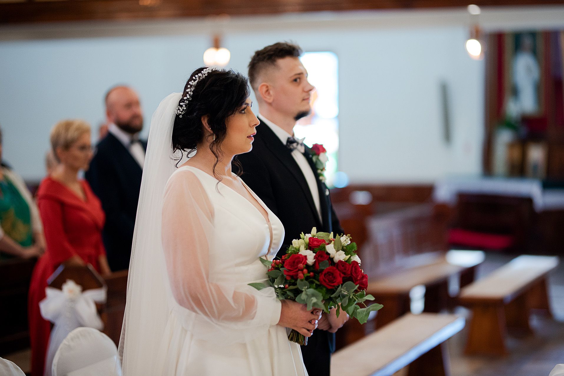 Para młoda podczas składania przysięgi małżeńskiej przy ołtarzu - panna młoda w białej sukni z tiulowymi rękawami i bukietem czerwonych róż, pan młody w czarnym garniturze