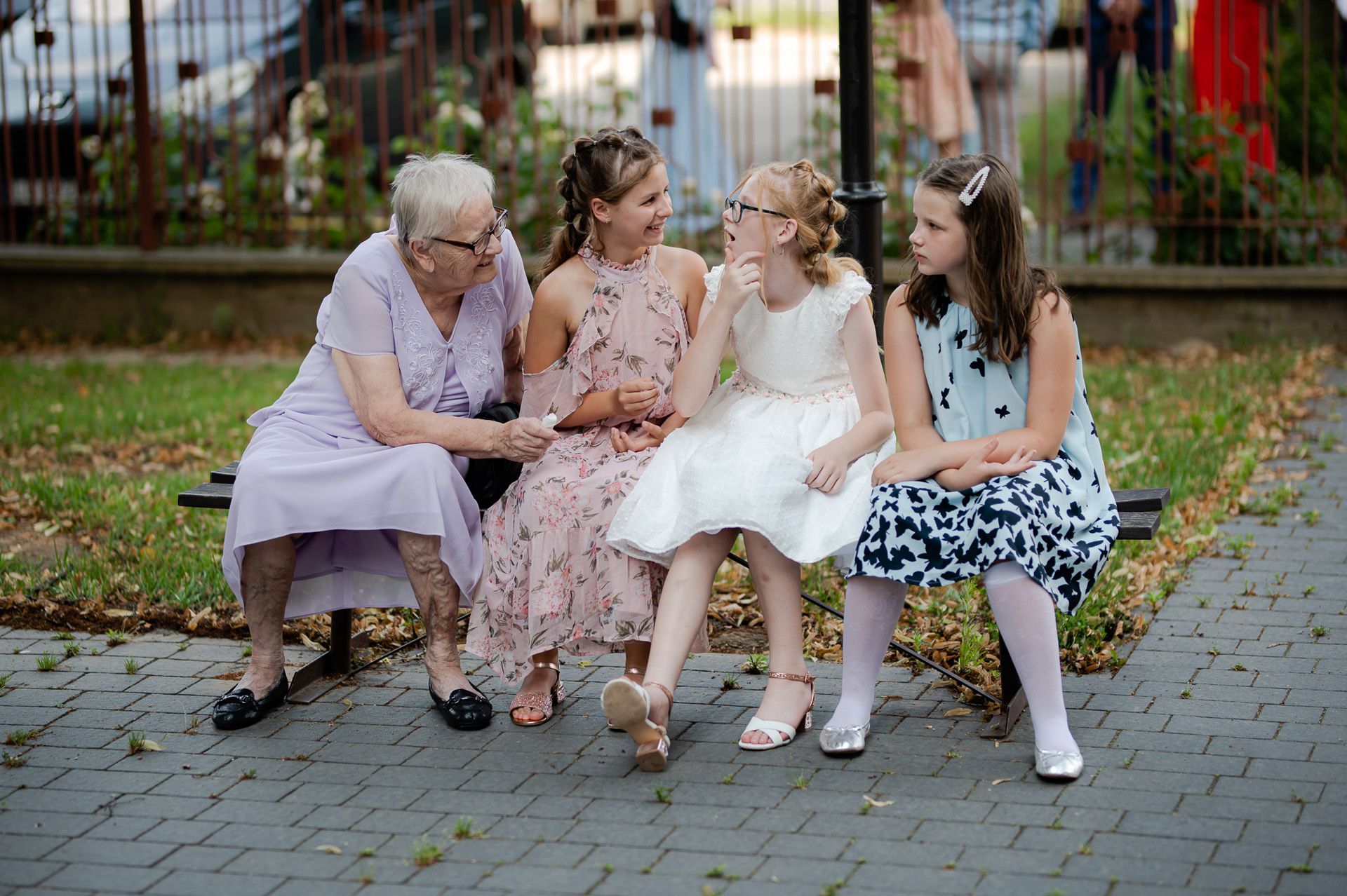 Wielopokoleniowa rodzina podczas ślubu - babcia z wnuczkami w eleganckich sukienkach siedzi na ławce w parku po ceremonii