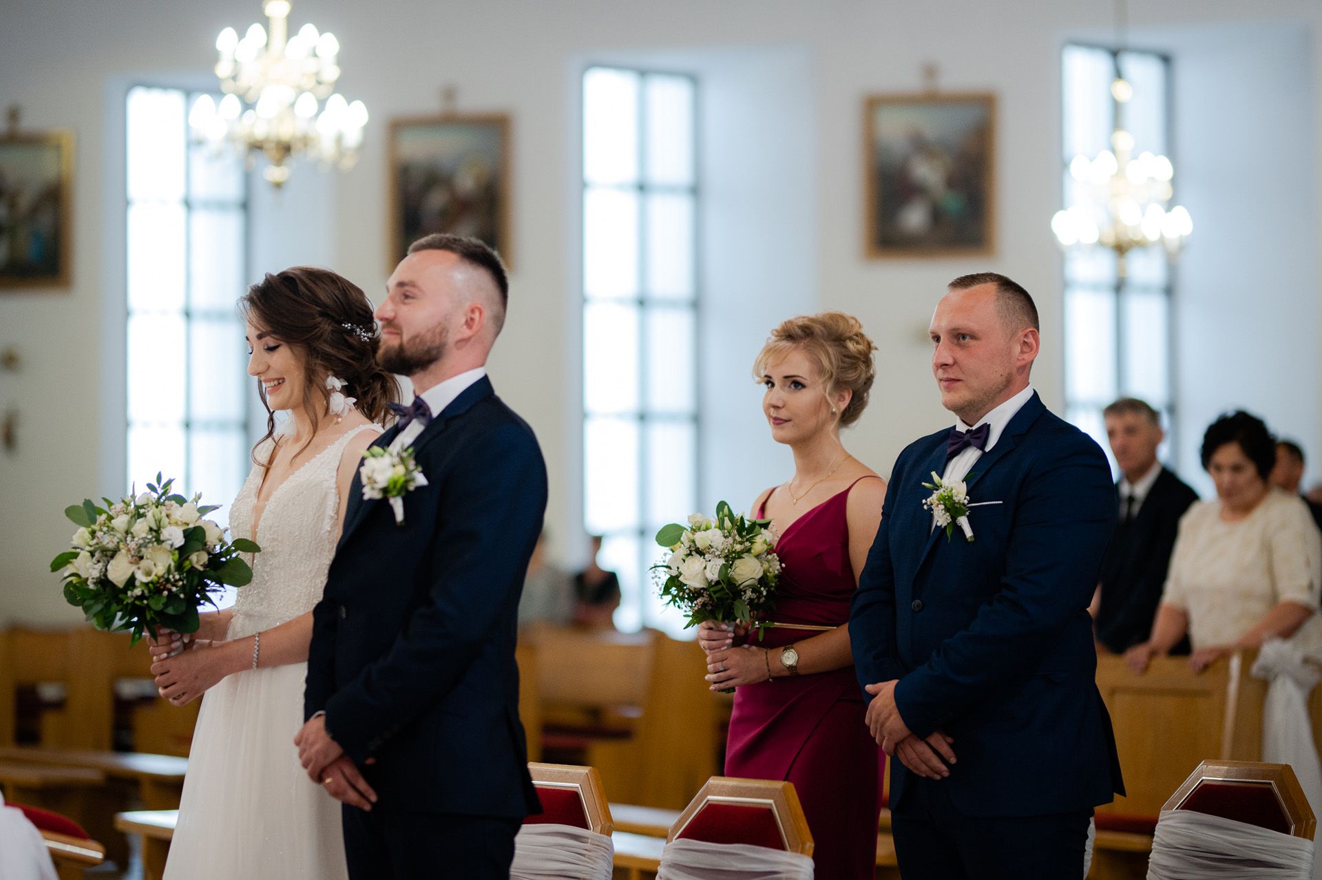 Ceremonia ślubu cywilnego w eleganckiej sali - para młoda ze świadkami w granatowych garniturach, druhny z bukietami, kryształowe żyrandole, białe wnętrze