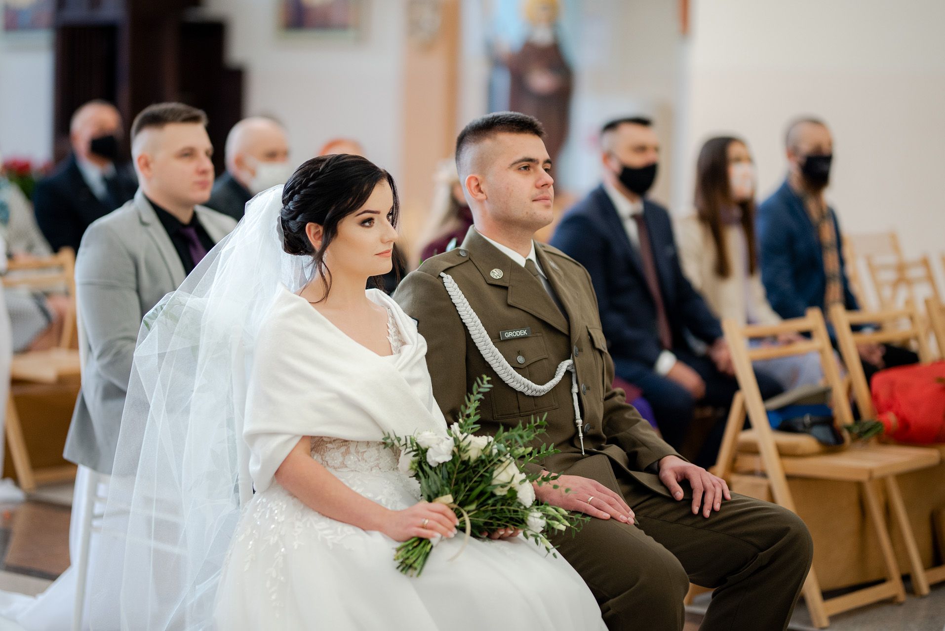 Para młoda podczas ceremonii ślubnej w kościele - panna młoda w białej sukni ślubnej z welonem i bukietem, pan młody w mundurze wojskowym, siedzą w pierwszej ławce