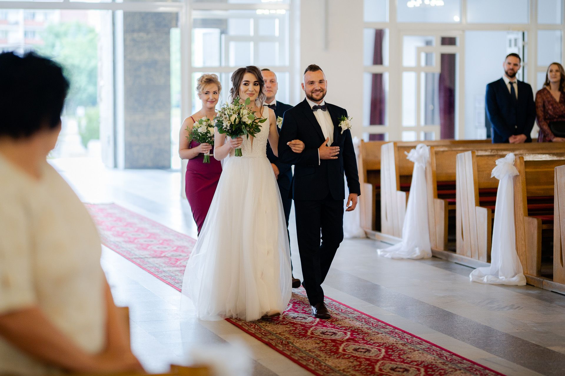 Para młoda wchodzi do kościoła czerwonym dywanem - panna młoda w białej sukni z bukietem, pan młody w czarnym smokingu, druhna w bordowej sukni, eleganckie wnętrze