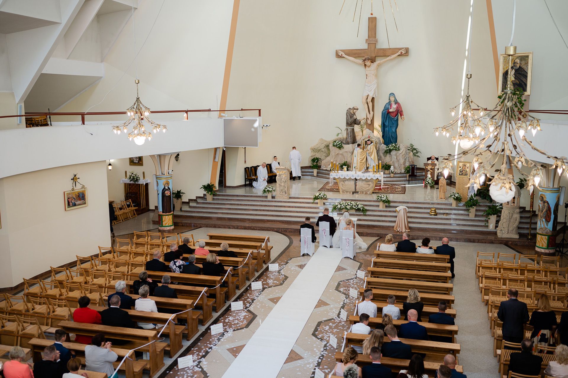 Panoramiczne ujęcie wnętrza kościoła podczas ceremonii ślubnej - widok z góry na ołtarz z krzyżem, ławki wypełnione gośćmi, białe dekoracje kwiatowe, para młoda przy ołtarzu