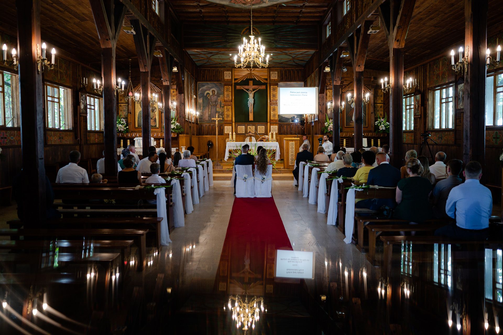 Wnętrze kościoła przygotowanego na ślub z czerwonym dywanem, białymi wstążkami na ławkach, goście oczekują na ceremonię