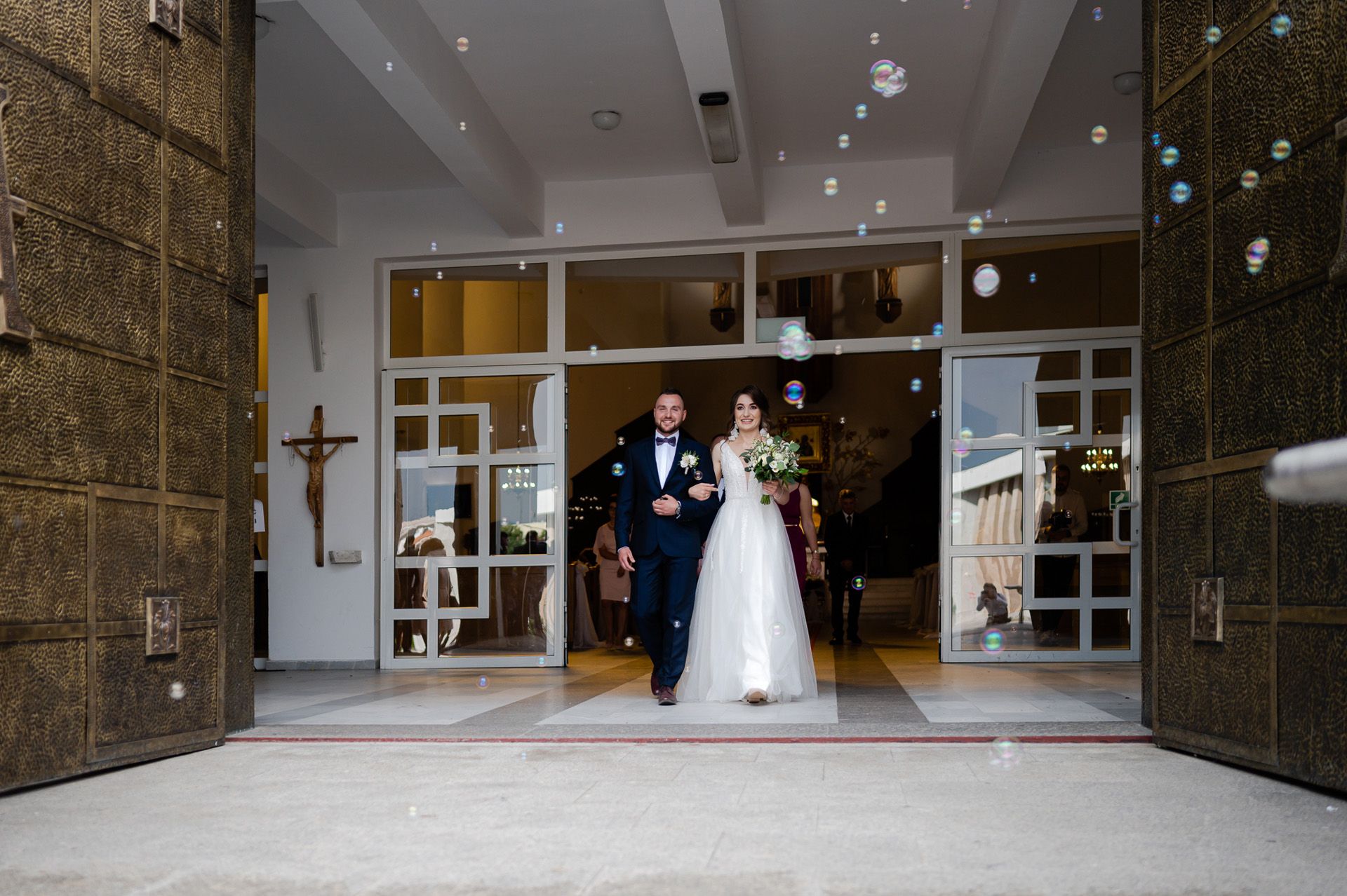 Para młoda wychodzi z nowoczesnego kościoła w otoczeniu baniek mydlanych - panna młoda w białej sukni z bukietem, pan młody w granatowym garniturze, współczesna architektura sakralna