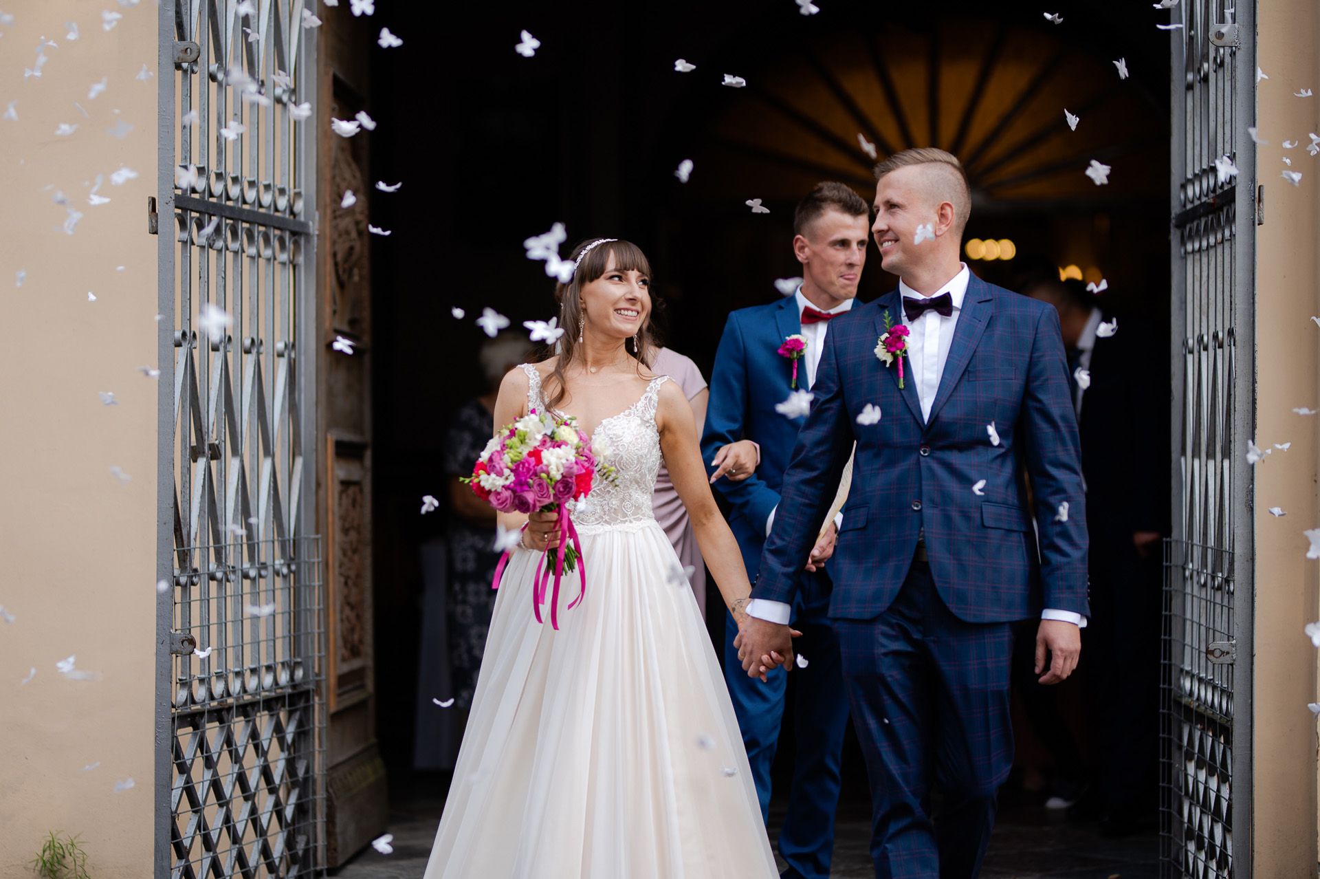 Para młoda wychodzi z kościoła w deszczu białego konfetti po ceremonii ślubnej - panna młoda w białej sukni z różowym bukietem, pan młody w granatowym garniturze, moment radości