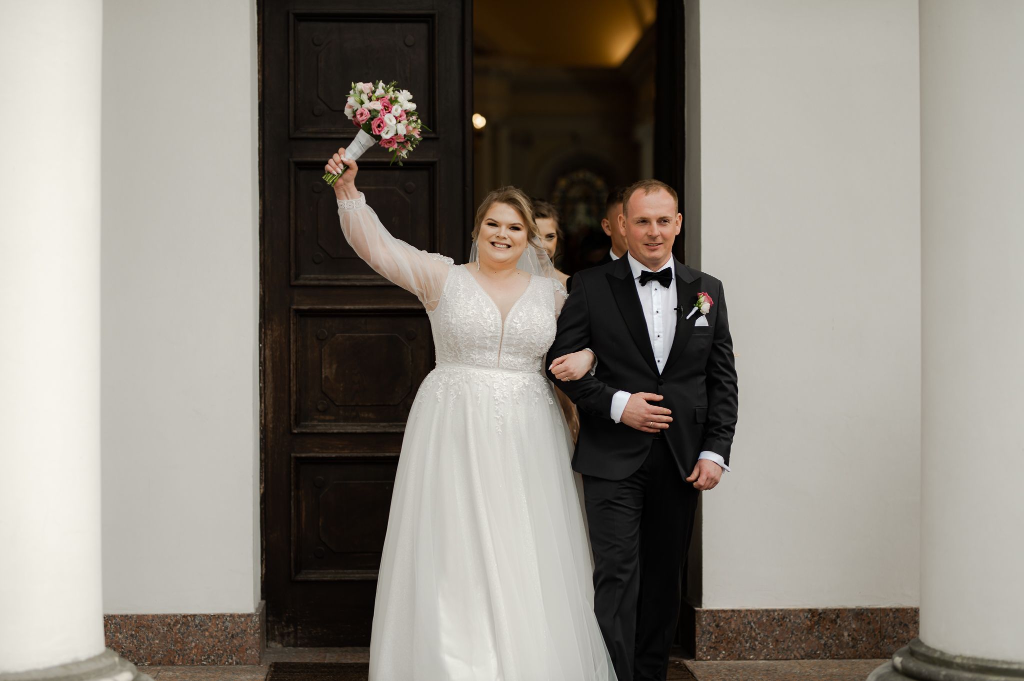 Para młoda wychodzi z kościoła po ceremonii ślubnej - panna młoda w białej sukni podnosi bukiet w geście radości, pan młody w czarnym garniturze z muszką