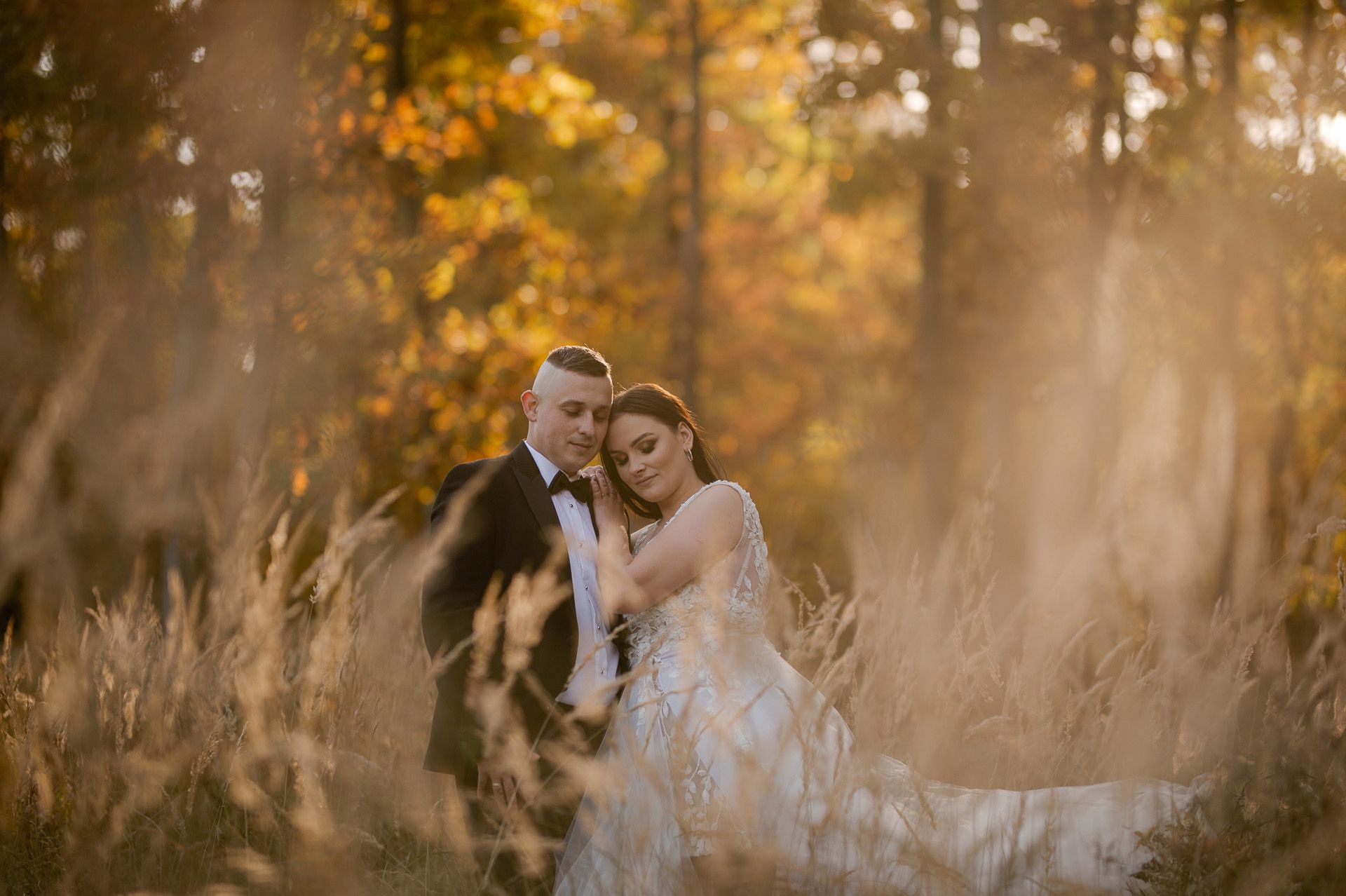 Czułe objęcie pary młodej wśród jesiennych traw - panna młoda z zamkniętymi oczami przytula się do pana młodego we fraku, miękkie światło słoneczne, romantyczna atmosfera sesji ślubnej