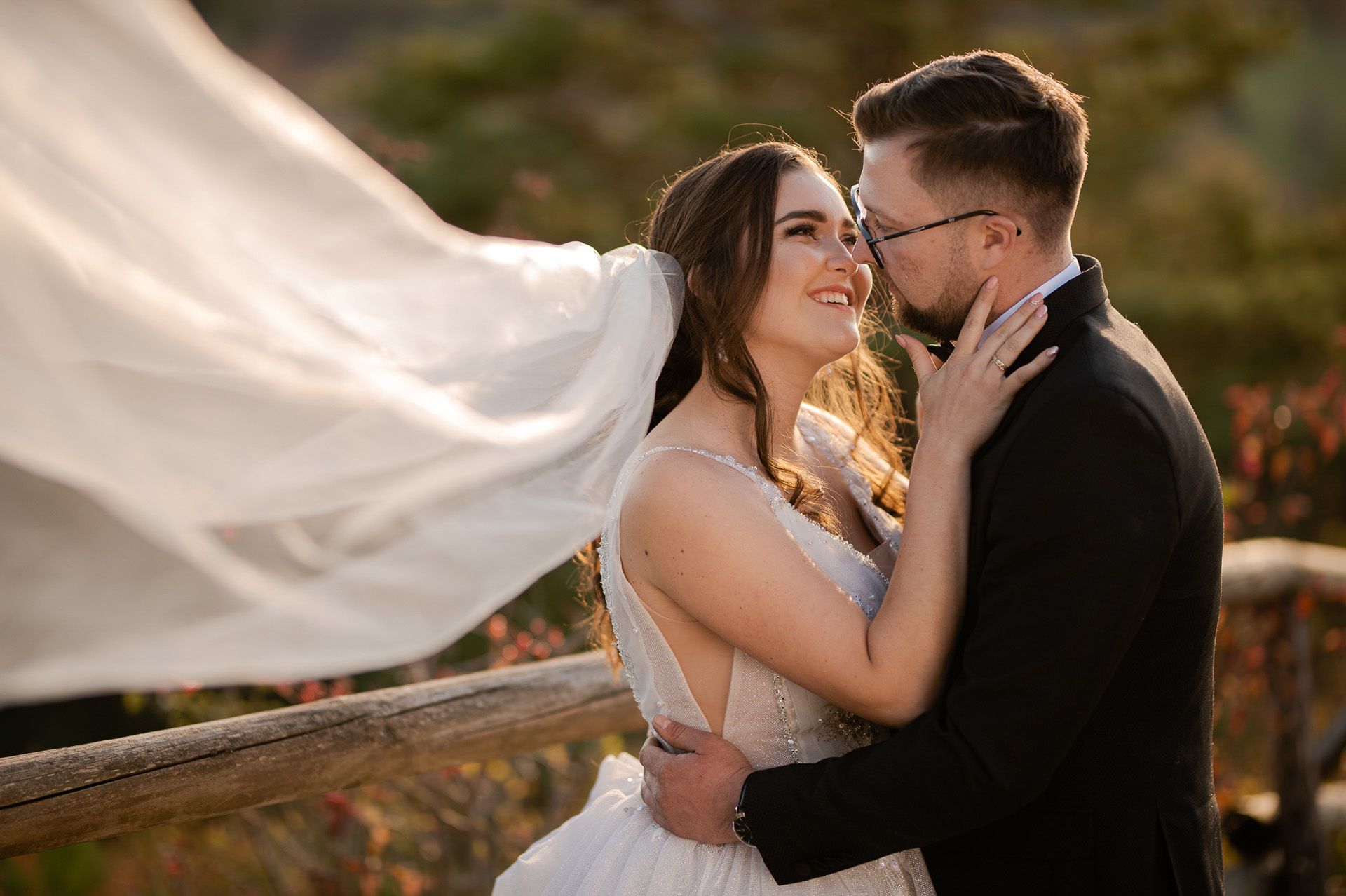 Intymna poza pary młodej podczas sesji ślubnej - panna młoda w sukni z perłowym zdobieniem i powiewającym welonem przytula się do pana młodego w czarnym garniturze, jesienne tło