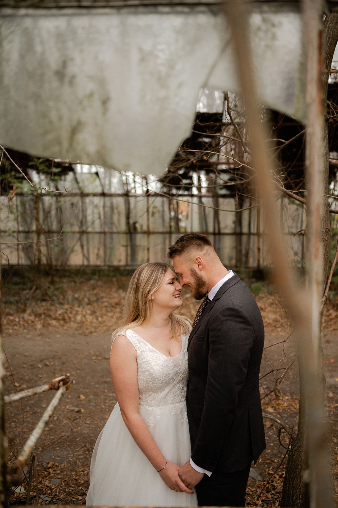 Intymny moment pary młodej na tle industrialnej architektury - blondynka panna młoda w prostej sukni ślubnej z koronkowym topem przytula się do pana młodego, surowe betonowe tło