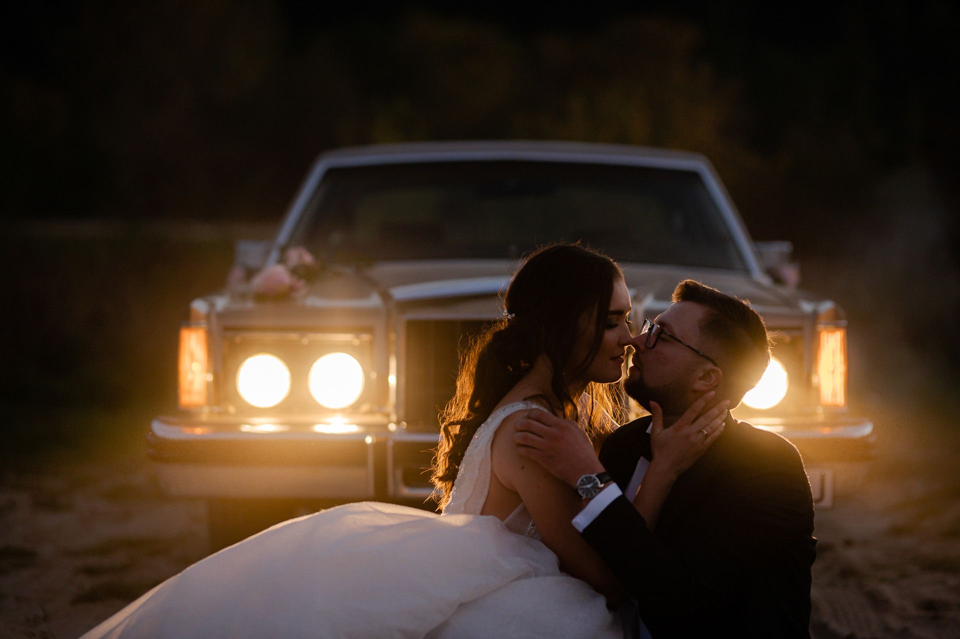 Nocna sesja ślubna pary młodej przy samochodzie z włączonymi reflektorami - romantyczny pocałunek w świetle reflektorów, panna młoda w białej sukni, pan młody w garniturze, klimatyczne oświetlenie