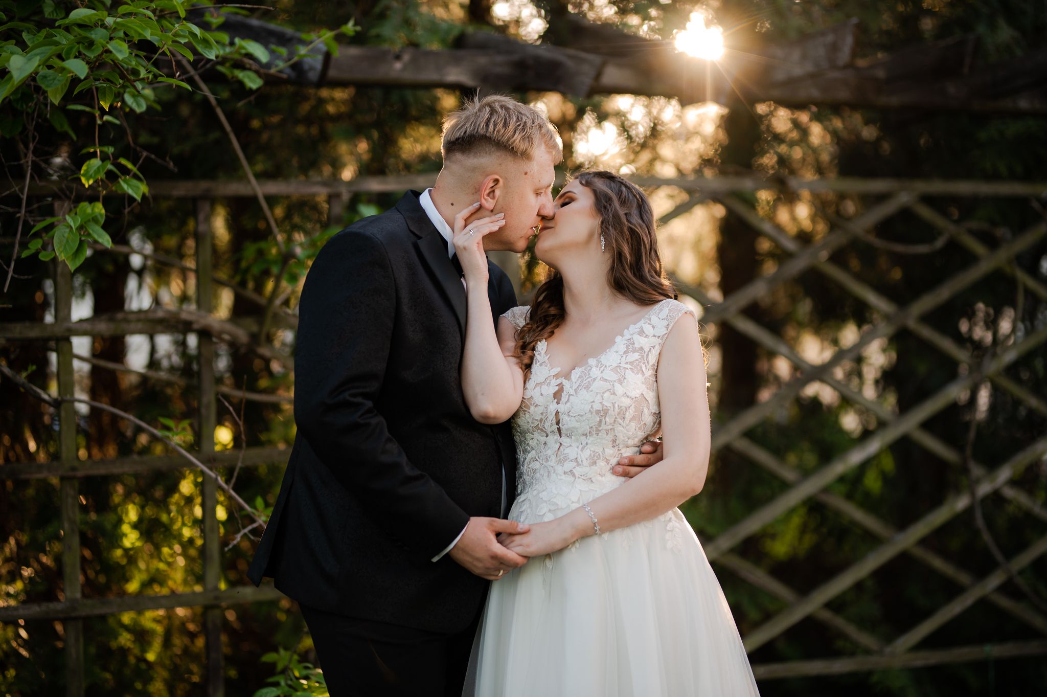 Romantyczny pocałunek pary młodej przy ogrodowej altanie - panna młoda w koronkowej sukni ślubnej całuje pana młodego w granatowym garniturze, słoneczne światło filtruje przez liście