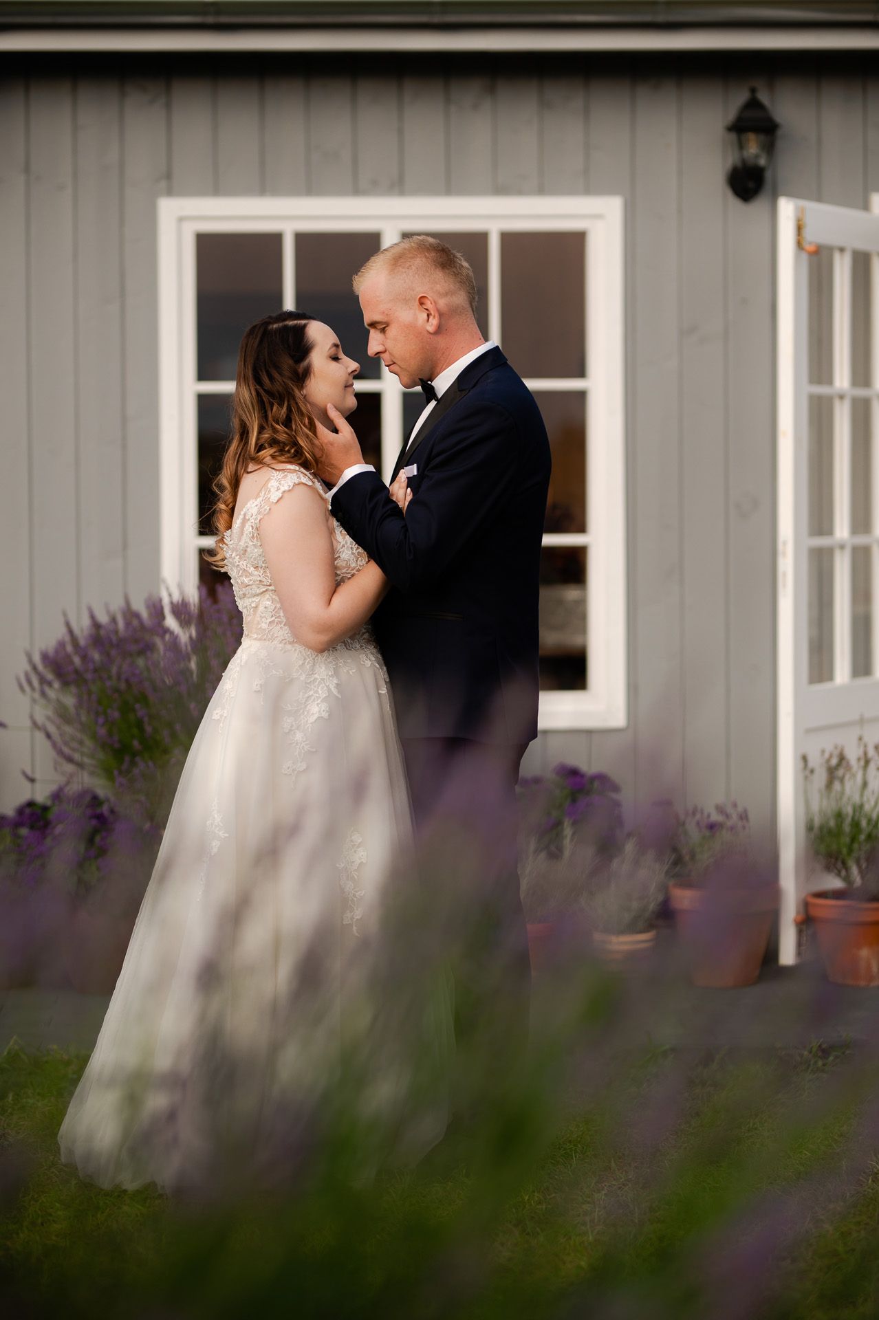 Romantyczna sesja ślubna przy wiejskim domu z lawendą - para młoda w czułym uścisku, panna młoda w koronkowej sukni bez rękawów, pan młody w granatowym garniturze, rustykalna sceneria