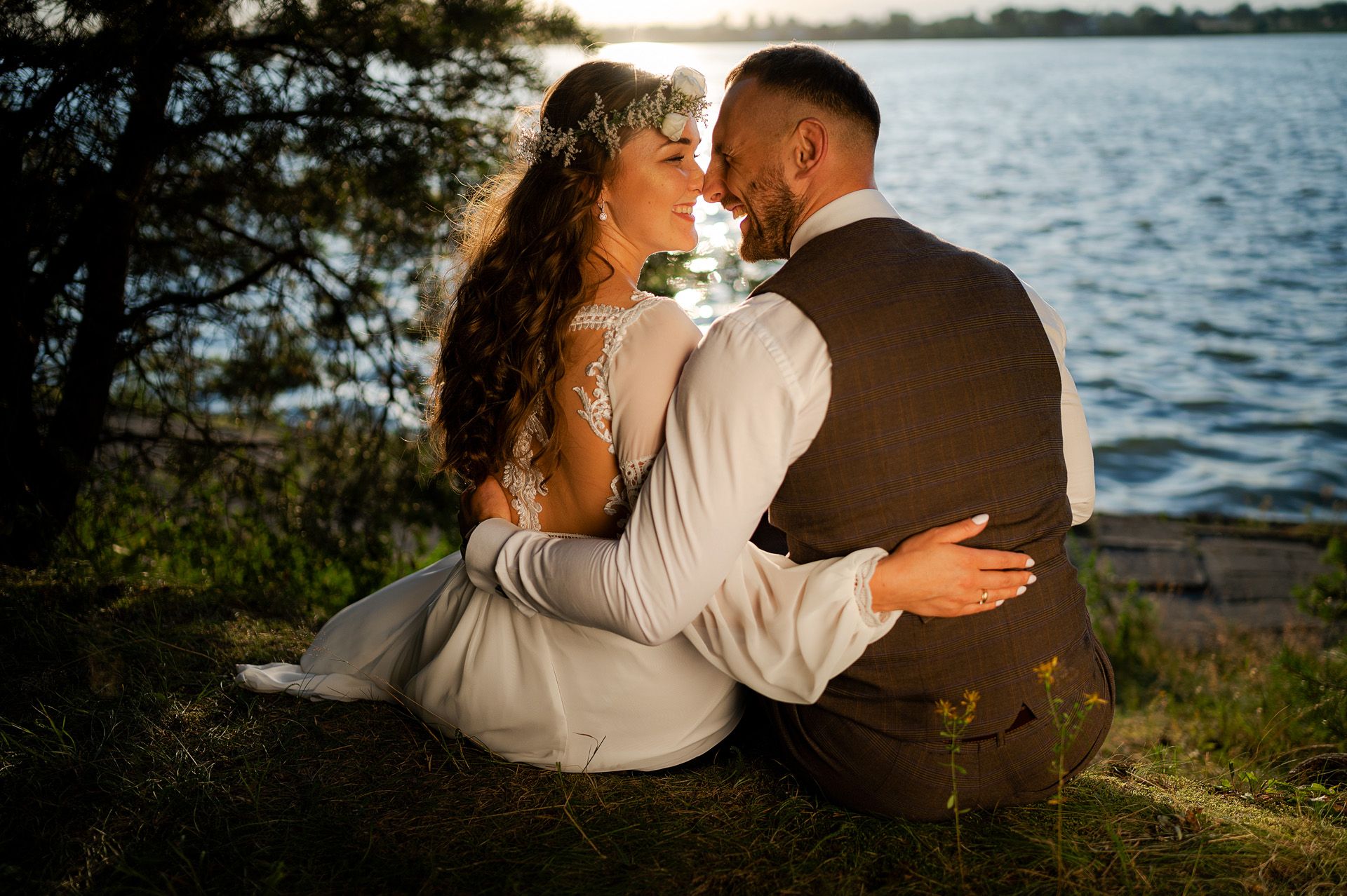 Sesja ślubna pary młodej nad jeziorem podczas zachodu słońca - panna młoda z wiankiem we włosach w koronkowej sukni ślubnej, pan młody w kamizelce, siedzą przytuleni na trawie, romantyczna atmosfera