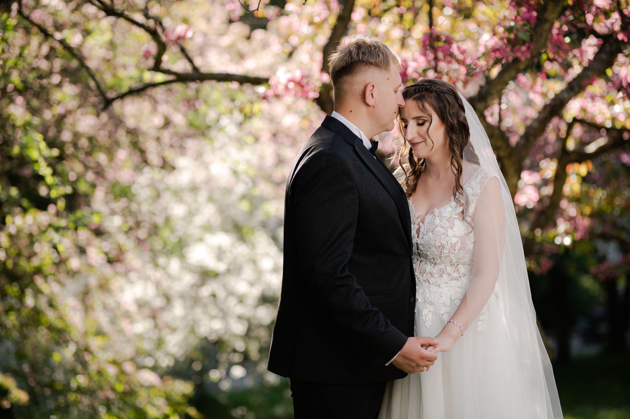 Sesja ślubna pod kwitnącym drzewem - panna młoda w długim welonie i koronkowej sukni ślubnej stoi blisko pana młodego w czarnym garniturze, różowe kwiaty na drzewie tworzą romantyczne tło