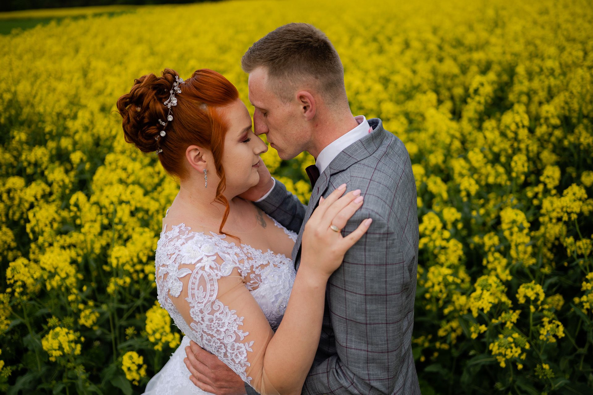 Para młoda podczas sesji ślubnej w kwitnącym polu rzepaku - panna młoda z rudymi włosami w koronkowej sukni ślubnej, pan młody w szarym garniturze, czułe objęcie wśród żółtych kwiatów