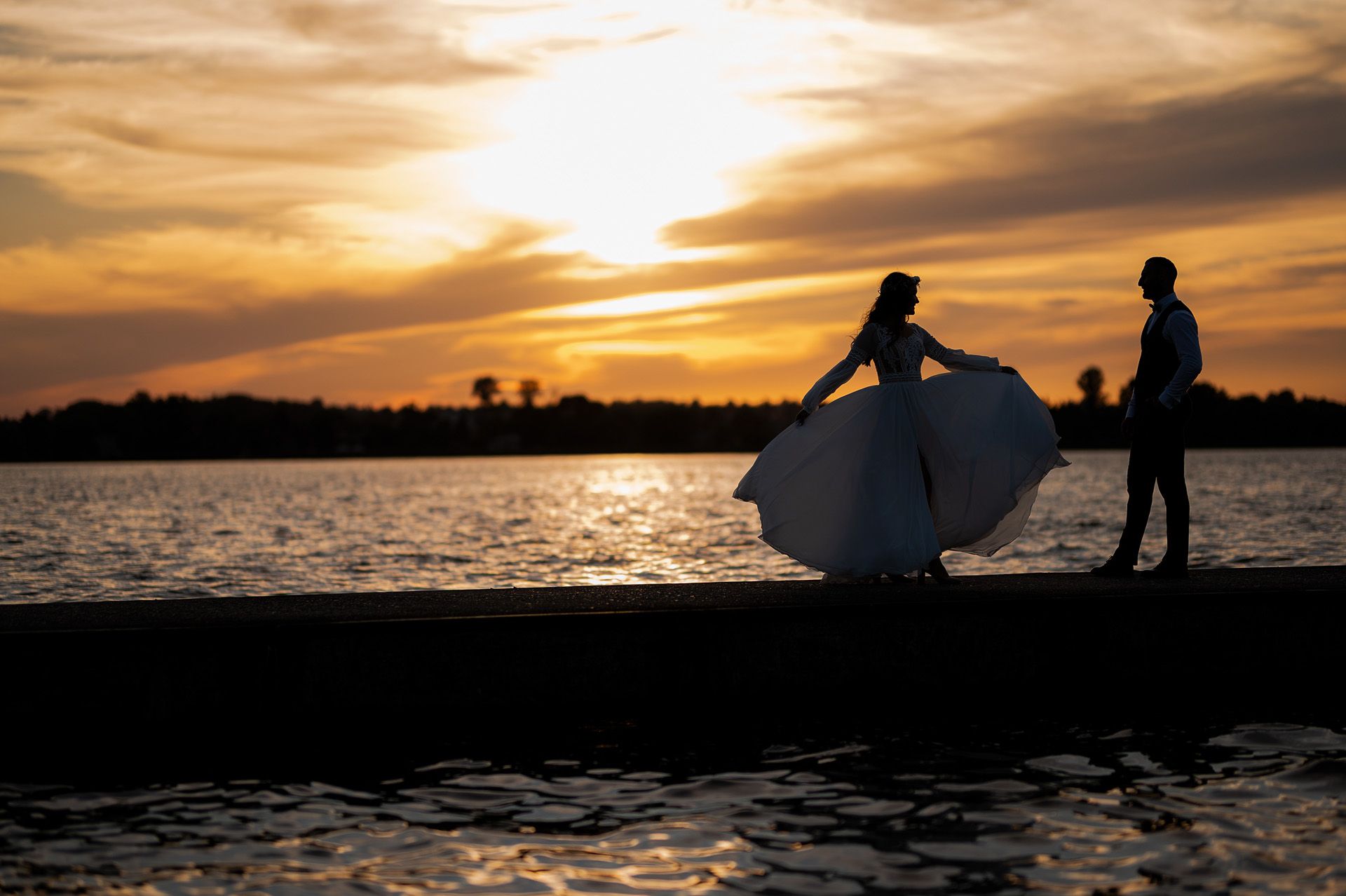 Sylwetki pary młodej podczas zachodu słońca nad jeziorem - panna młoda w pływającej sukni ślubnej tańczy z panem młodym, złote niebo i odbicia w wodzie, artystyczna fotografia ślubna