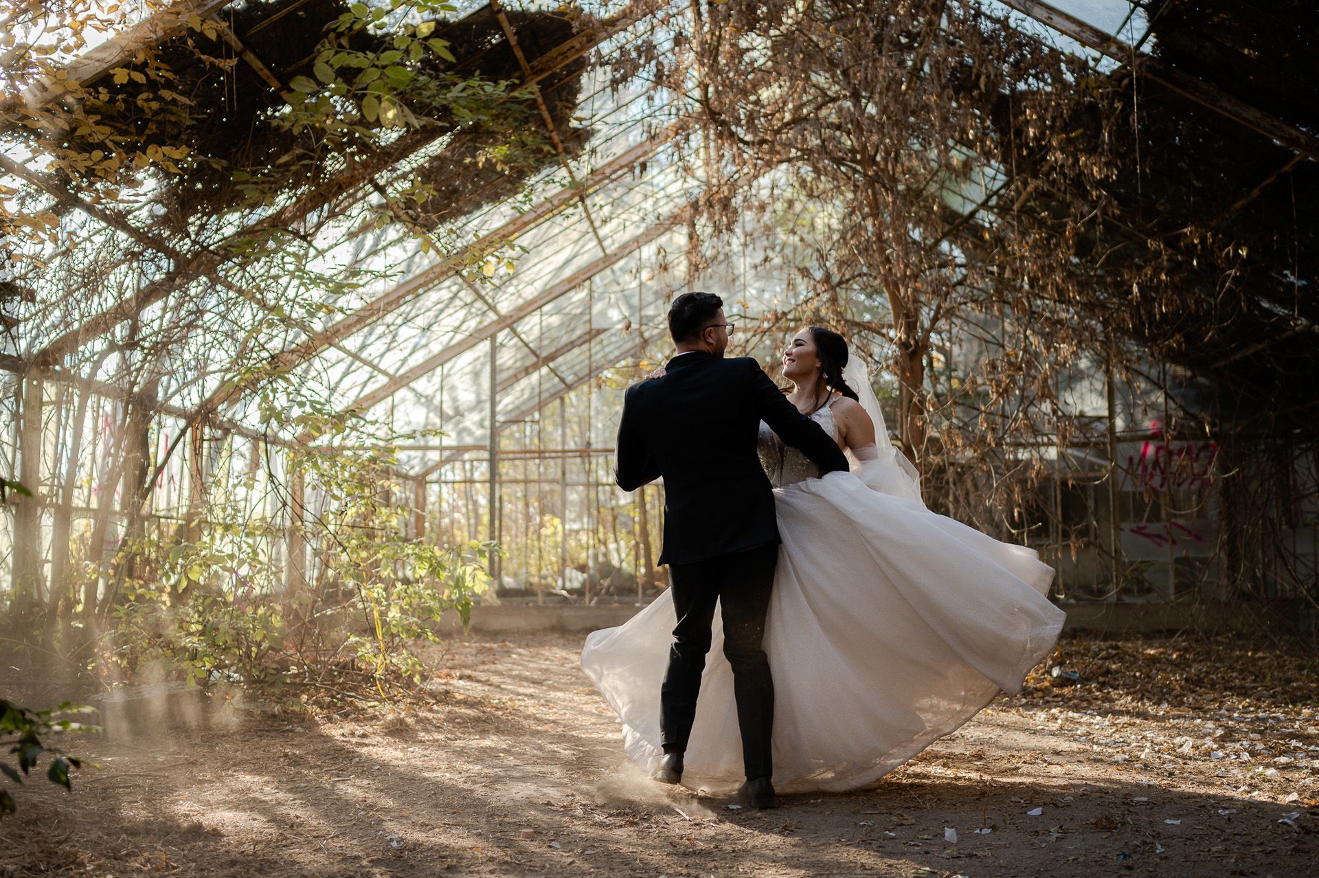 Romantyczny taniec pary młodej w opuszczonej szklarni - pan młody prowadzi pannę młodą w wirującym tańcu, suknia ślubna unosi się w powietrzu, magiczna atmosfera z naturalnym światłem