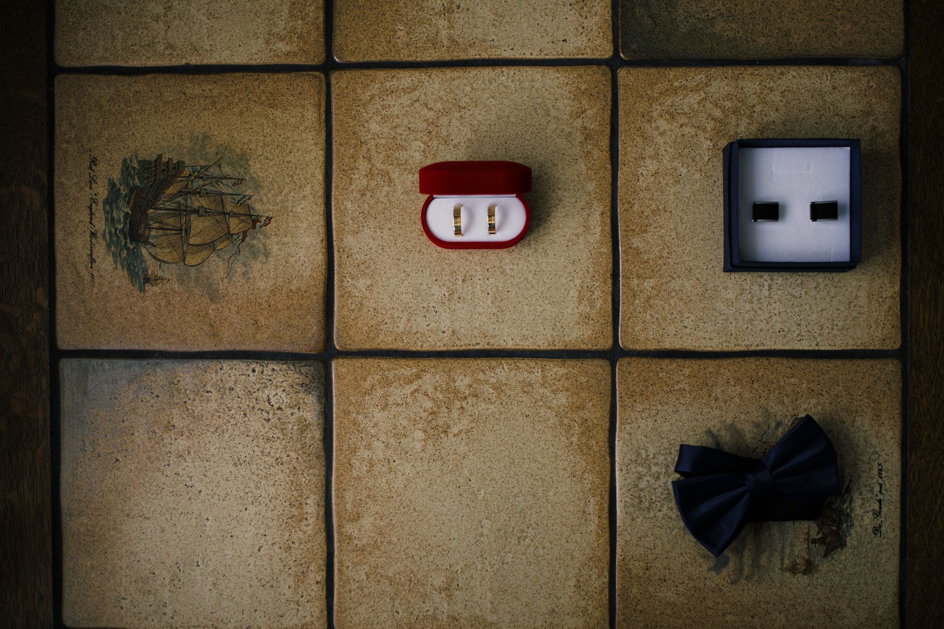 Złote obrączki ślubne w czerwonym pudełku położone na kamiennych kaflach obok spinek do mankietów i muszki