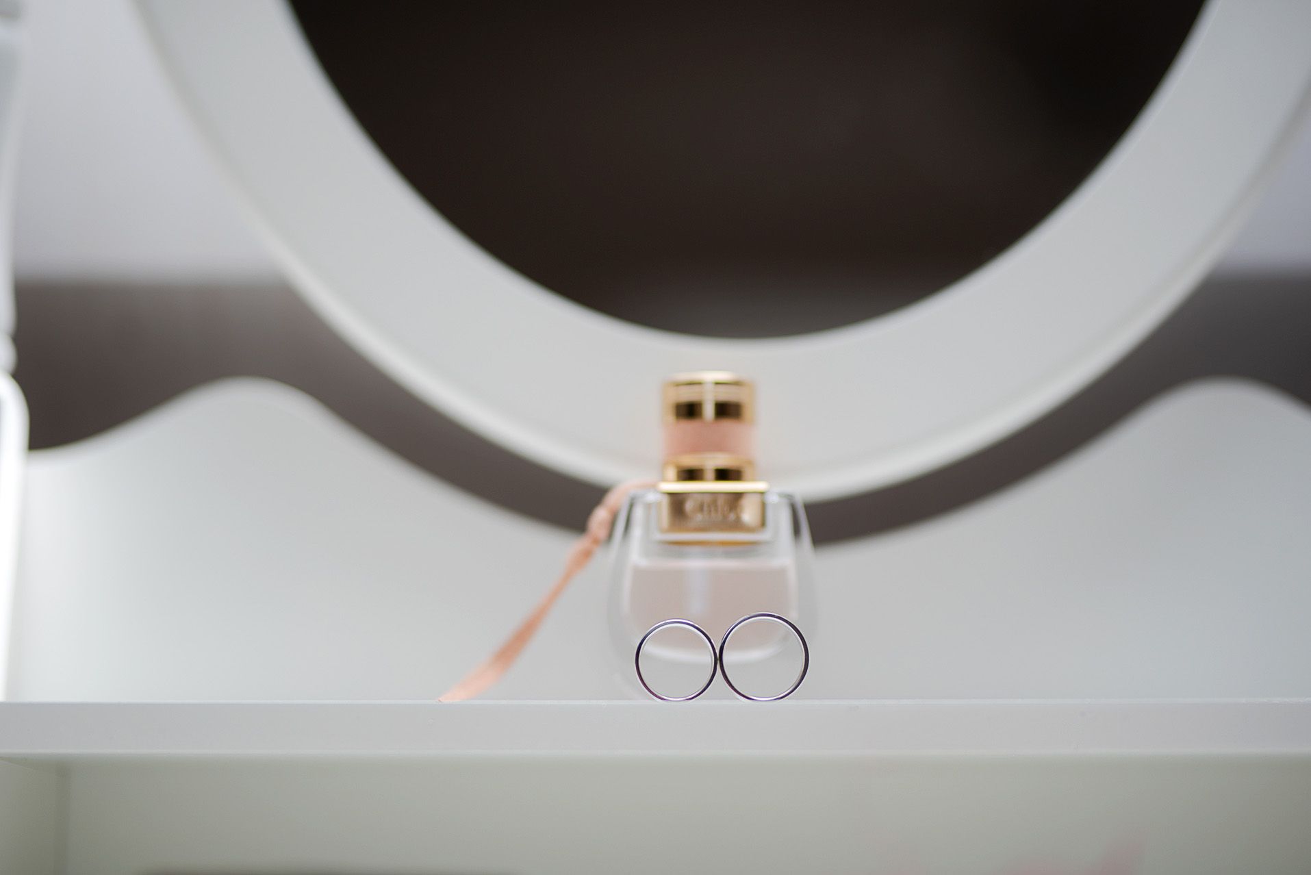 Złote obrączki ślubne i perfumy ułożone na białej toaletce przy okrągłym lustrze