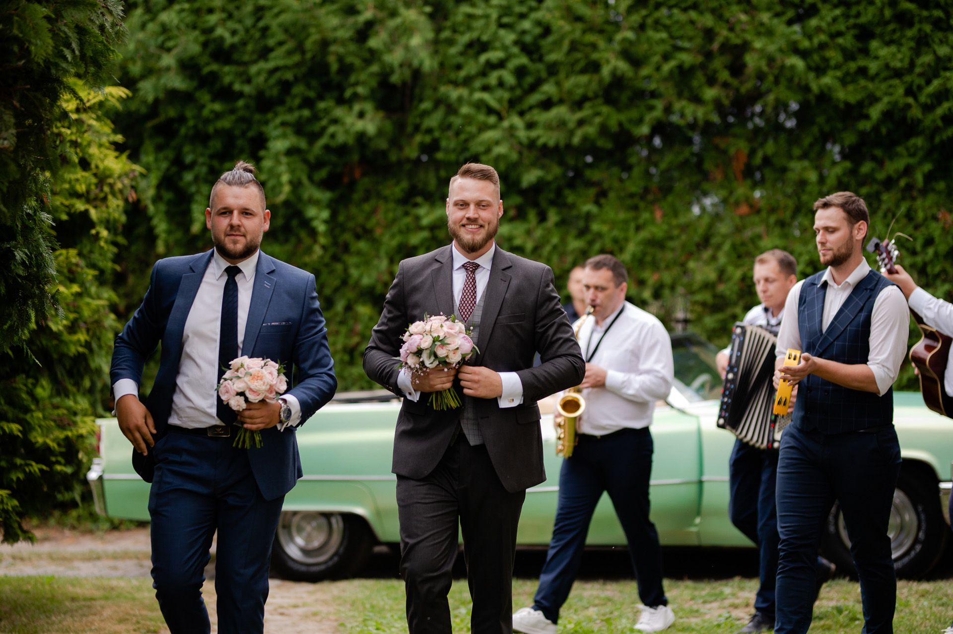 Pan młody z druhami w garniturach trzymający bukiety na tle zielonego zabytkowego samochodu z zespołem muzycznym