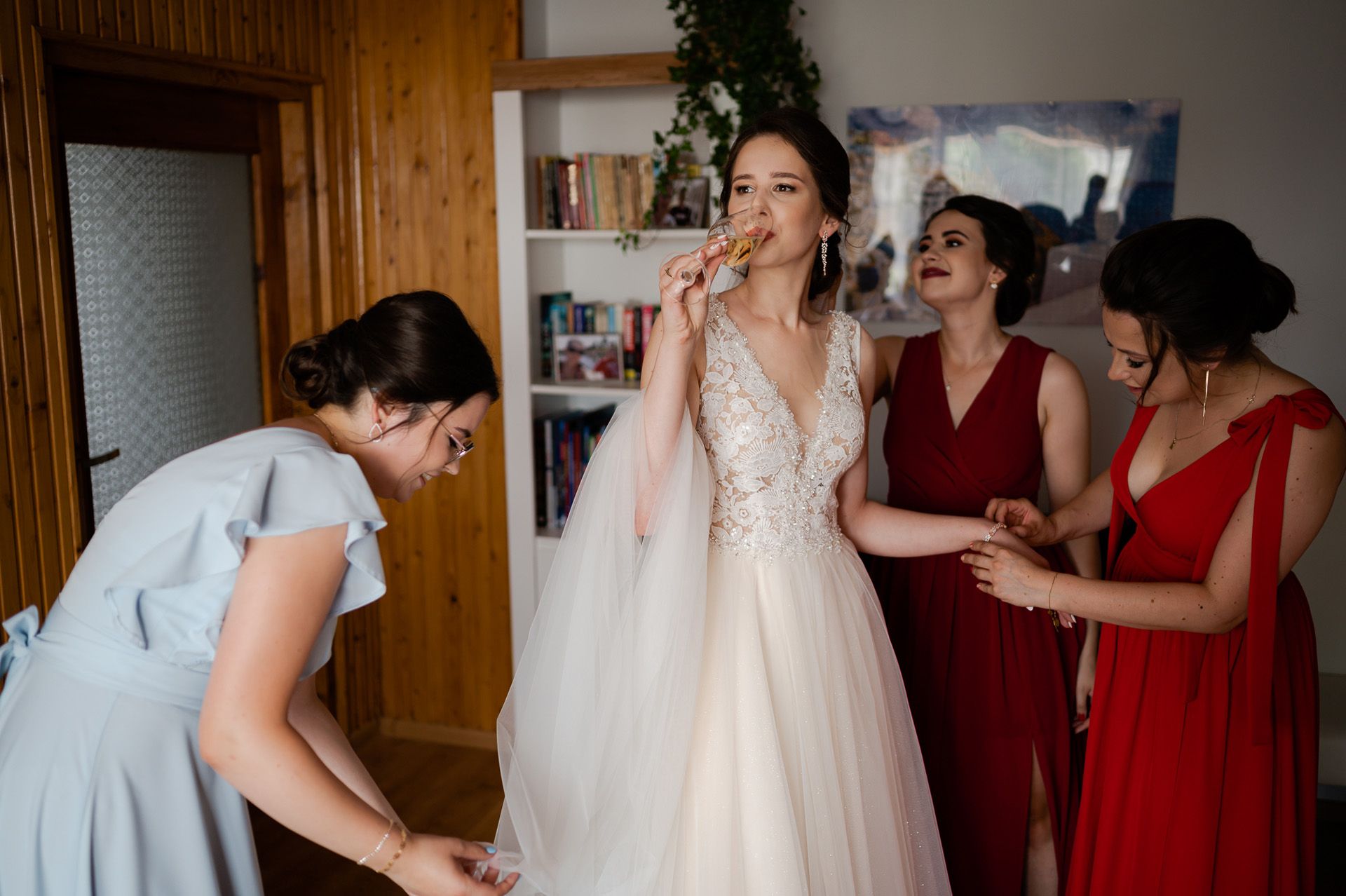 Panna młoda w koronkowej sukni z welonem pijąca szampana otoczona druhnami w czerwonych sukniach