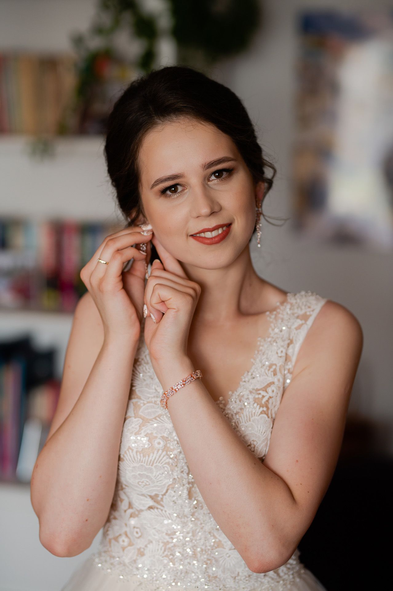 Uśmiechnięta panna młoda w koronkowej sukni zakładająca perłowe kolczyki przed ślubem