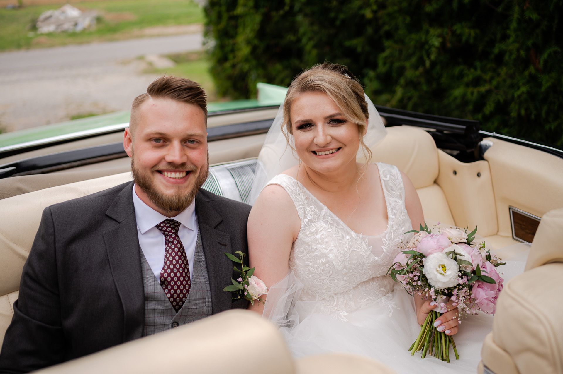 Szczęśliwa para młoda siedząca w jasnym kabriolecie, panna młoda z bukietem różowo-białym