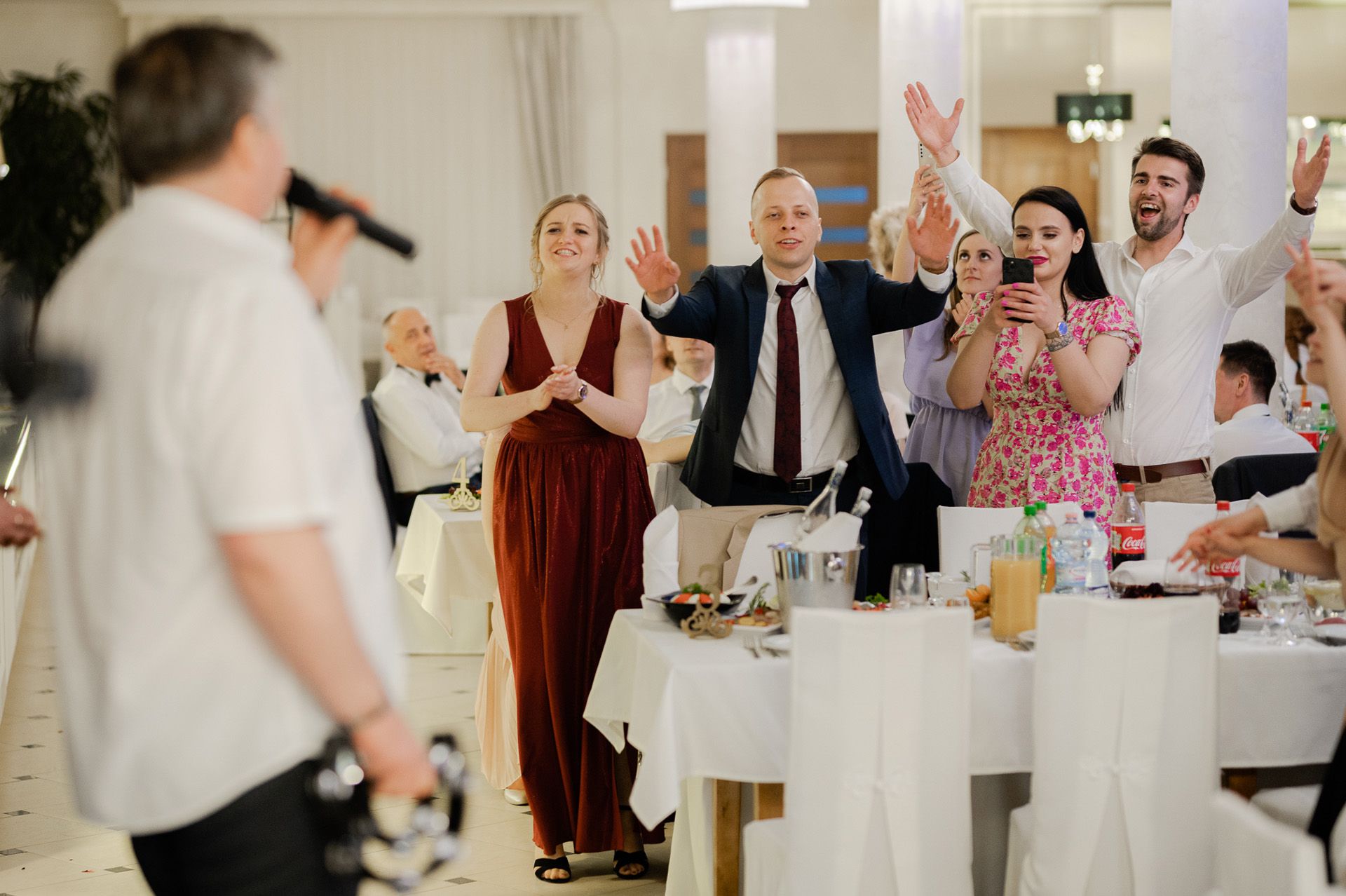 Goście weselni bawią się podczas animacji w sali weselnej, podnoszą ręce do góry, kobieta w bordowej sukni i mężczyzna w granatowym garniturze, radosna atmosfera przyjęcia ślubnego