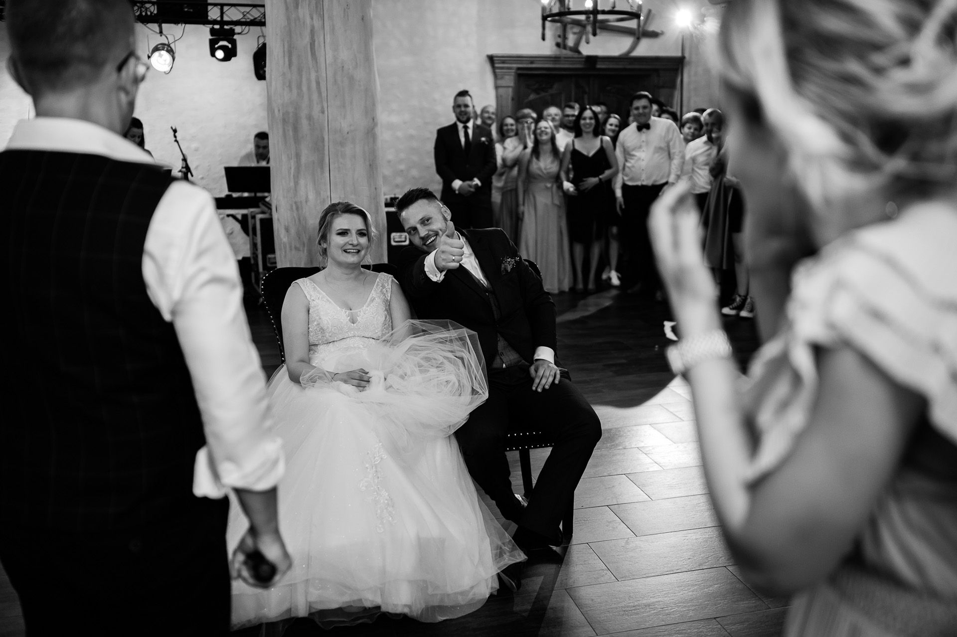 Para młoda siedzi podczas toastu weselnego, panna młoda w tiulowej sukni, pan młody wskazuje na gości, czarno-białe zdjęcie