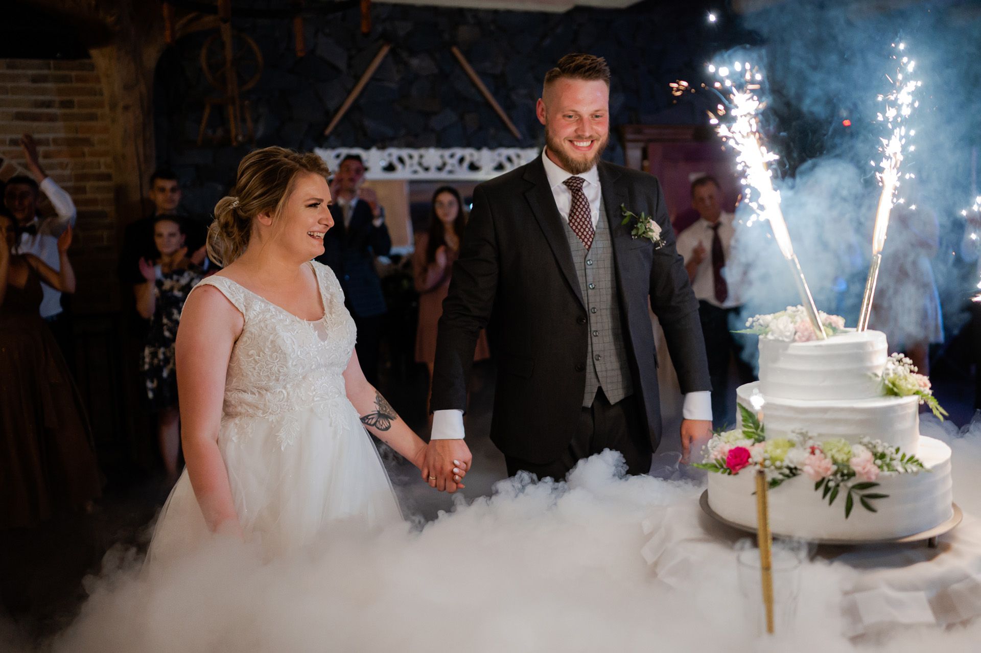 Para młoda przy torcie ślubnym z zimnymi ogniami w rustykalnym wnętrzu, biały tort z kwiatami, ceremonia krojenia tortu