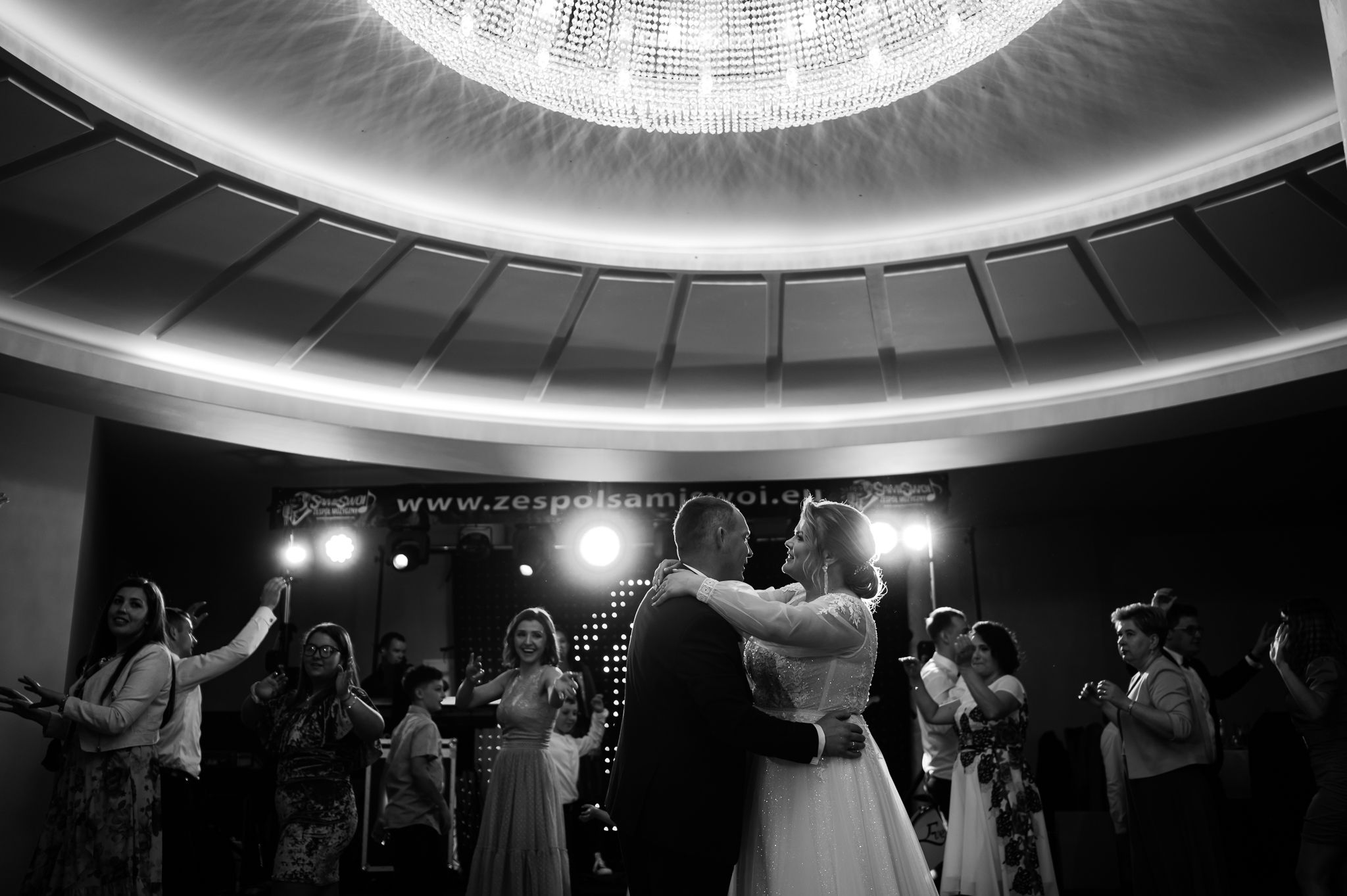 Pierwszy taniec pary młodej w eleganckiej sali weselnej z okrągłym sufitem i kryształowym żyrandolem, czarno-białe zdjęcie ślubne
