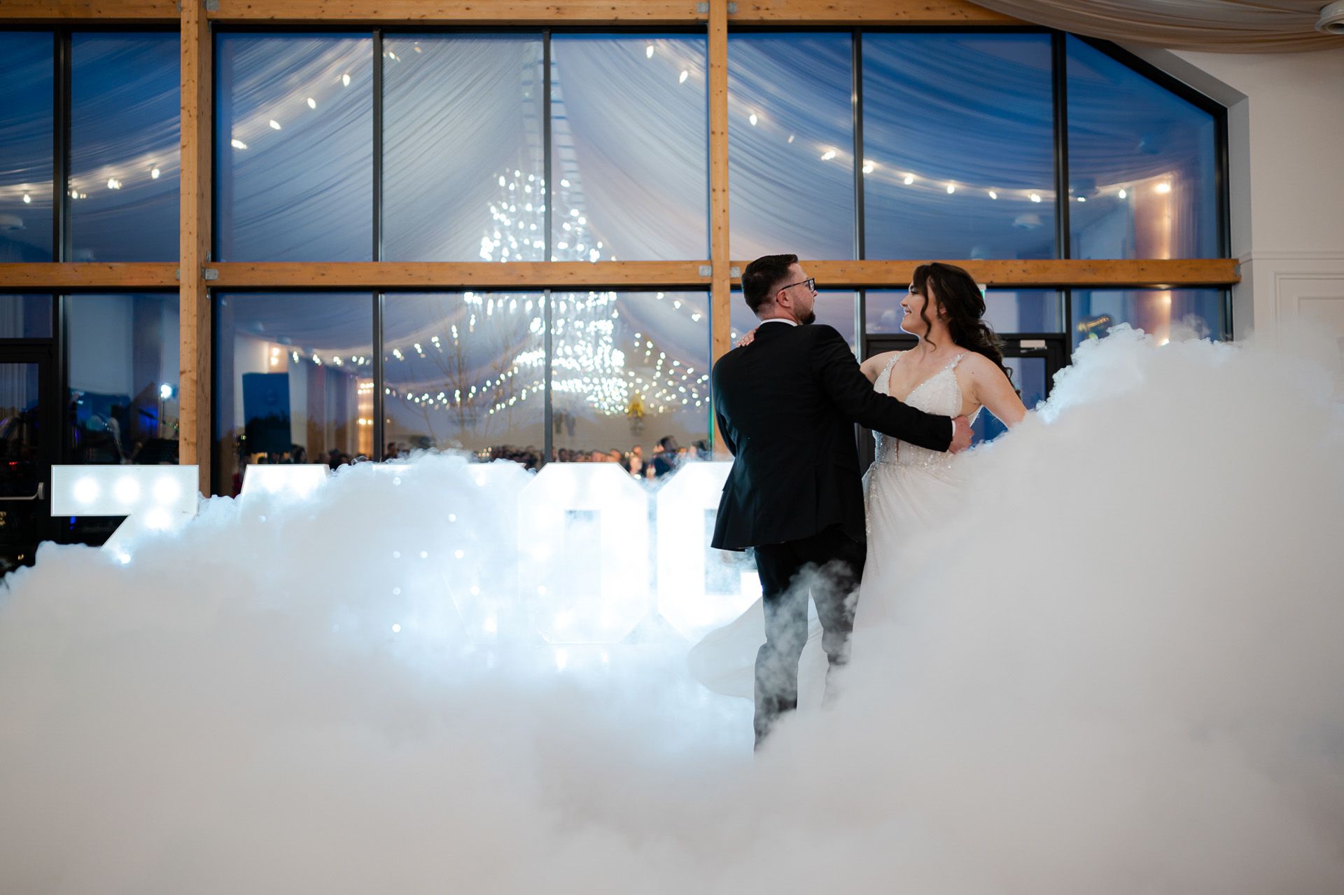 Para młoda tańczy romantyczny taniec w chmurze dymu ze świecącymi lampkami w tle, panna młoda w białej sukni ślubnej, pan młody w czarnym garniturze