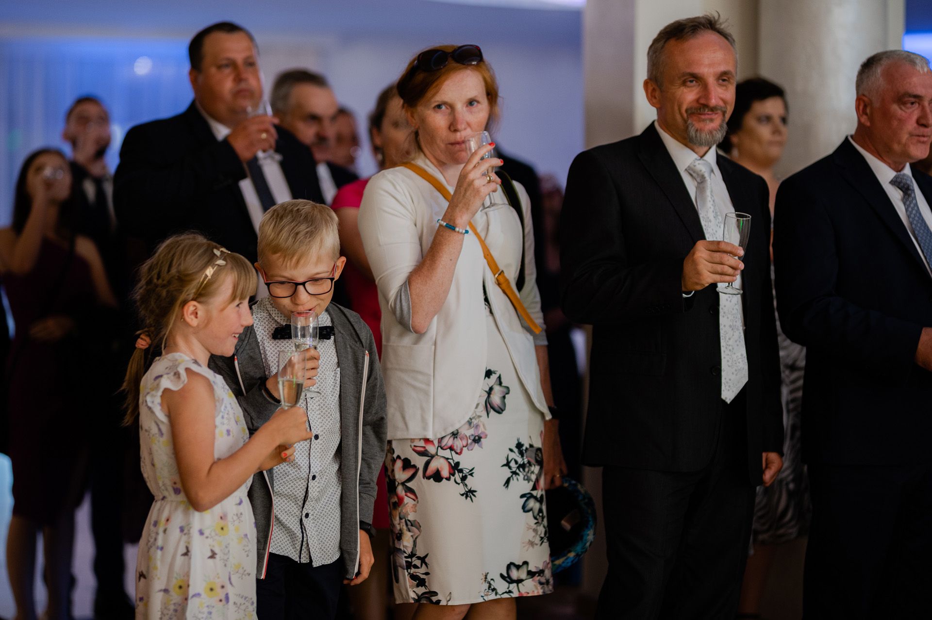 Toast weselny z udziałem całej rodziny, dzieci z kieliszkami, goście wznoszą toast za młodą parę
