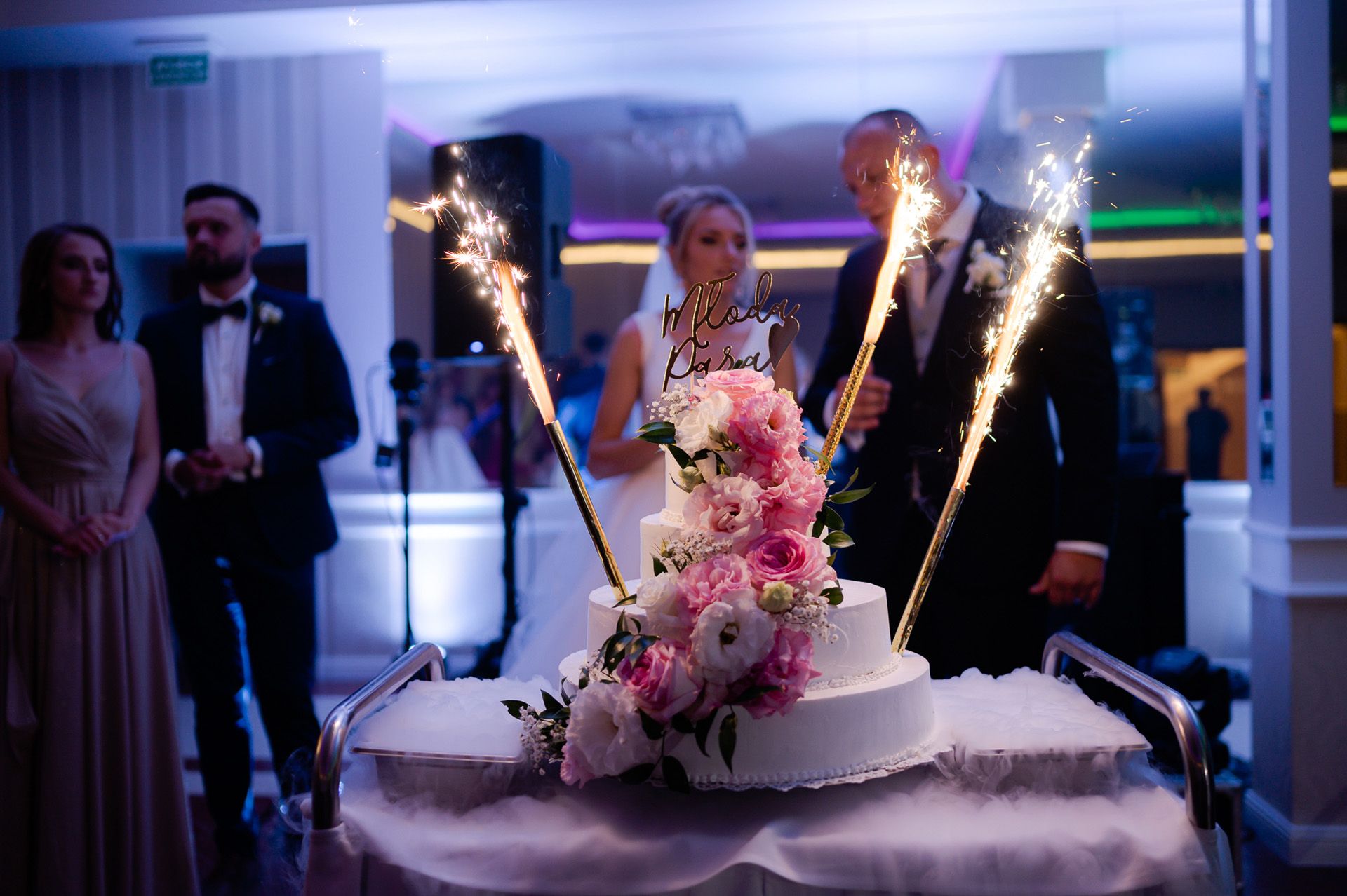 Para młoda kroi tort weselny z zimnymi ogniami, biały tort z różowymi kwiatami, romantyczny moment ceremonii ślubnej